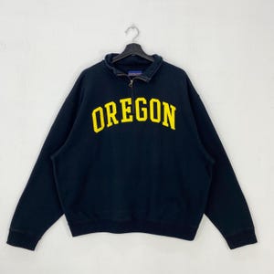 May include: Black sweatshirt with a quarter-zip collar and the word "OREGON" in yellow block letters across the chest. The sweatshirt has long sleeves and a ribbed collar and cuffs. The garment is hanging on a black hanger.