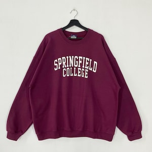 Puede incluir: Una sudadera burdeos de Springfield College con letras blancas. La sudadera de cuello redondo está colgada de una percha negra sobre un fondo blanco. La parte superior de manga larga está hecha de un material suave.