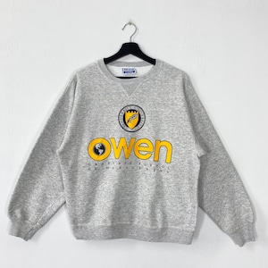 Könnte beinhalten: Ein meliertes graues Sweatshirt mit dem Wort "Owen" in gelben Blockbuchstaben. Das Sweatshirt hat auch ein rundes Emblem und den Text "Graduate School of Management". Das Sweatshirt hängt an einem schwarzen Kleiderbügel.