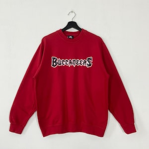 Puede incluir: Una sudadera roja de manga larga con cuello redondo que presenta la palabra Buccaneers bordada en el pecho con una tipografía estilizada en blanco y negro. La prenda tiene cuello, puños y dobladillo acanalados y se muestra en una percha negra.