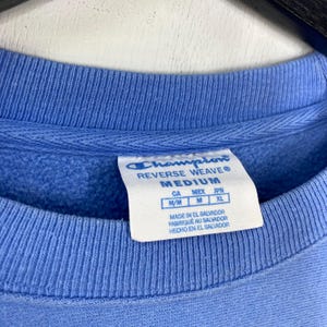 Peut inclure: Gros plan d'un sweat-shirt Champion Reverse Weave bleu clair. L'&eacute;tiquette indique une taille moyenne, avec des mesures pour CA, MEX et JPN. L'&eacute;tiquette indique &eacute;galement que l'article a &eacute;t&eacute; fabriqu&eacute; au Salvador.