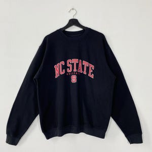 May include: A black NC State Wolfpack sweatshirt with the words "NC STATE" in red and white arched across the chest. Below the text is the word "WOLFPACK" and a red "S" logo. The sweatshirt is hanging on a black hanger.