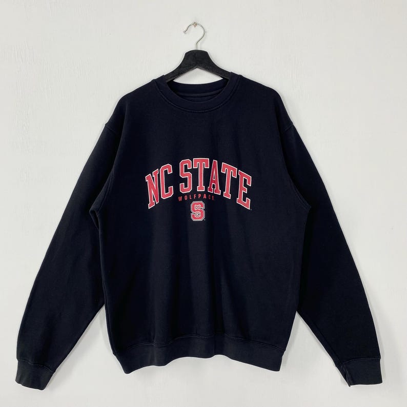 May include: A black NC State Wolfpack sweatshirt with the words "NC STATE" in red and white arched across the chest. Below the text is the word "WOLFPACK" and a red "S" logo. The sweatshirt is hanging on a black hanger.