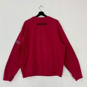 K&ouml;nnte beinhalten: Ein rotes Sweatshirt mit dem Text "WOLFPACK" in Schwarz auf dem oberen R&uuml;cken gedruckt. Auf dem linken &Auml;rmel steht das Wort "STATE" in Wei&szlig;. Das Sweatshirt h&auml;ngt an einem schwarzen Kleiderb&uuml;gel.