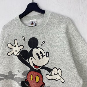 Może przedstawiać: Jasnoszara bluza z dużym nadrukiem Myszki Miki. Myszka Miki jest przedstawiona w swoich klasycznych kolorach: czarnym, czerwonym i ż&oacute;łtym. Bluza wisi na czarnym wieszaku.