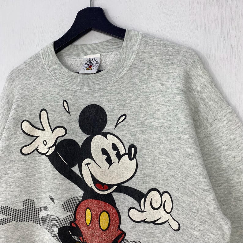 Może przedstawiać: Jasnoszara bluza z dużym nadrukiem Myszki Miki. Myszka Miki jest przedstawiona w swoich klasycznych kolorach: czarnym, czerwonym i ż&oacute;łtym. Bluza wisi na czarnym wieszaku.