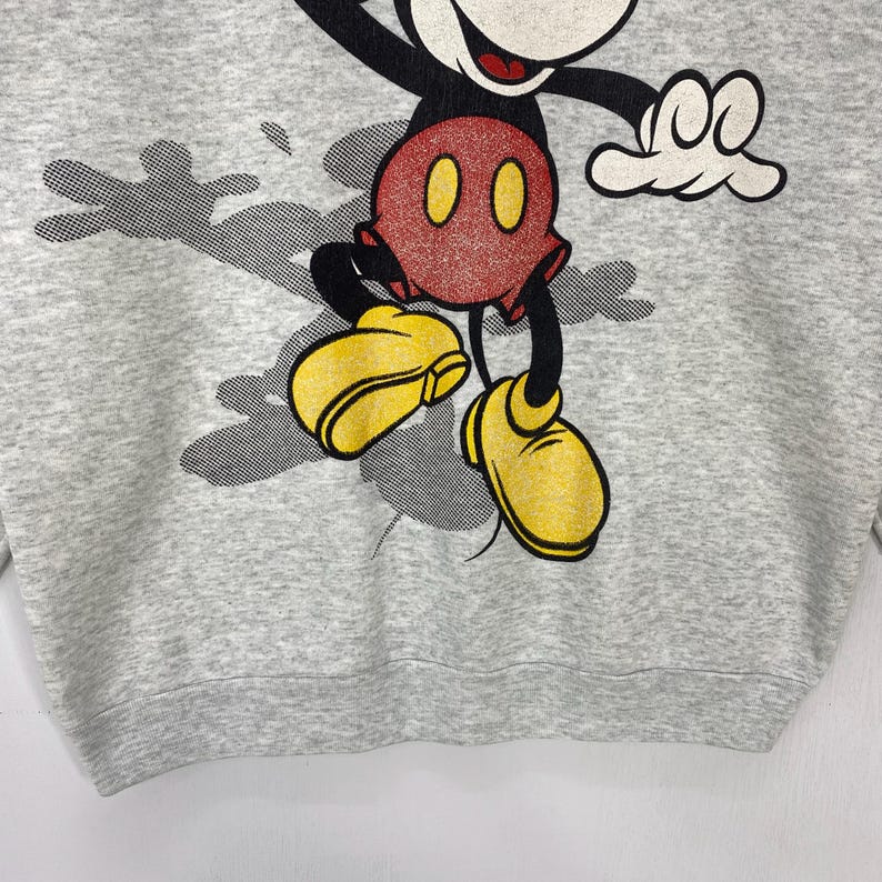 Może przedstawiać: Szara bluza z dużym nadrukiem Myszki Miki. Postać z kresk&oacute;wki jest przedstawiona w kolorach czarnym, czerwonym i ż&oacute;łtym, w dynamicznej pozie. Bluza ma okrągły dekolt i długie rękawy.