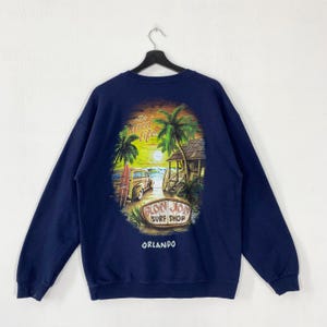 Op de afbeelding: Marineblauwe sweatshirt met een kleurrijke afbeelding van een strandscène, inclusief palmbomen, een surfshop en een vintage truck. De tekst "Ron Jon Surf Shop Orlando" staat eronder.