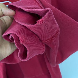 K&ouml;nnte beinhalten: Nahaufnahme eines karmesinroten Sweatshirt-&Auml;rmels. Das gerippte B&uuml;ndchen weist Gebrauchsspuren mit kleinen Rissen auf. Der Stoff ist dick und scheint aus einem weichen Material zu bestehen. Die N&auml;hte sind entlang der N&auml;hte sichtbar.