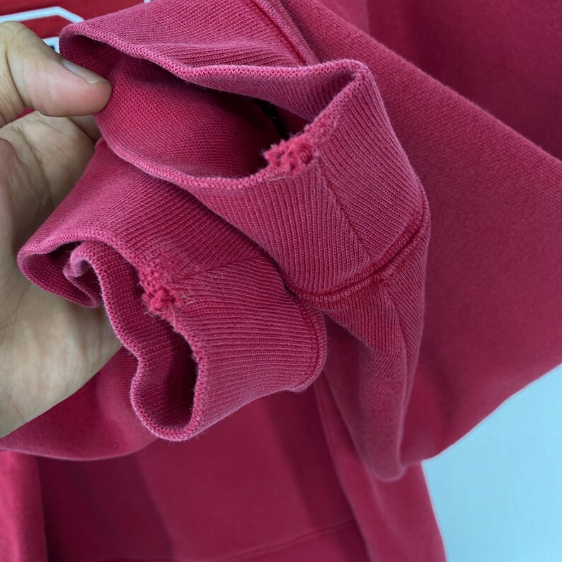 K&ouml;nnte beinhalten: Nahaufnahme eines karmesinroten Sweatshirt-&Auml;rmels. Das gerippte B&uuml;ndchen weist Gebrauchsspuren mit kleinen Rissen auf. Der Stoff ist dick und scheint aus einem weichen Material zu bestehen. Die N&auml;hte sind entlang der N&auml;hte sichtbar.