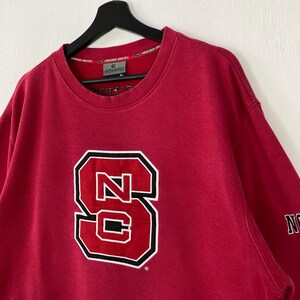 K&ouml;nnte beinhalten: Ein rotes Sweatshirt mit einem gro&szlig;en, wei&szlig; umrandeten, schwarz umrandeten "NC"-Logo. Das Logo ist mittig auf der Vorderseite platziert. Das Sweatshirt hat einen Rundhalsausschnitt und lange &Auml;rmel. Der Markenname "Colosseum Athletics" ist auf dem Kragen aufgedruckt.