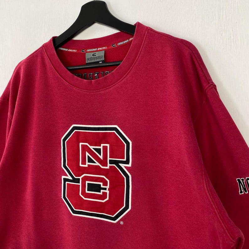 K&ouml;nnte beinhalten: Ein rotes Sweatshirt mit einem gro&szlig;en, wei&szlig; umrandeten, schwarz umrandeten "NC"-Logo. Das Logo ist mittig auf der Vorderseite platziert. Das Sweatshirt hat einen Rundhalsausschnitt und lange &Auml;rmel. Der Markenname "Colosseum Athletics" ist auf dem Kragen aufgedruckt.