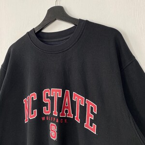 May include: Black crew neck sweatshirt with the words "NC STATE" in red block letters with a white outline. The word "WOLFPACK" is below the word "STATE". The sweatshirt is hanging on a black hanger.