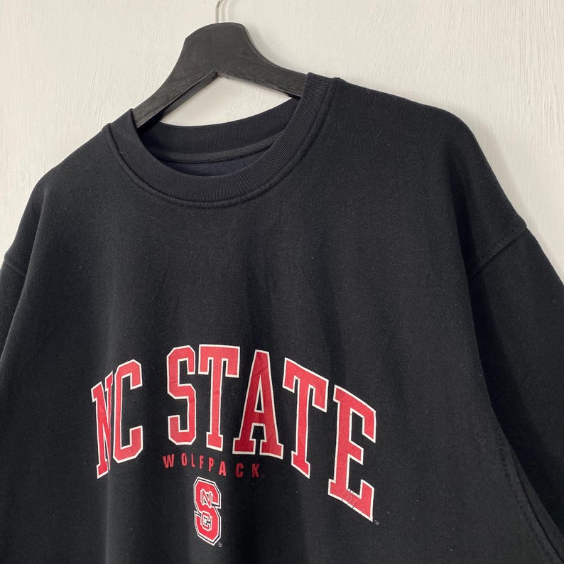 May include: Black crew neck sweatshirt with the words "NC STATE" in red block letters with a white outline. The word "WOLFPACK" is below the word "STATE". The sweatshirt is hanging on a black hanger.