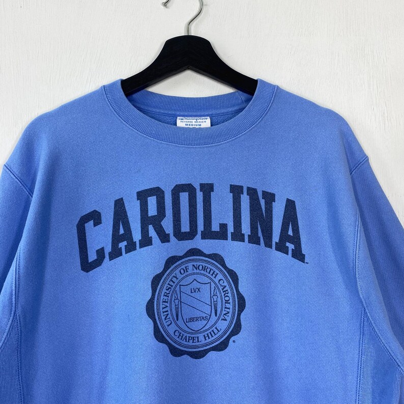 Peut inclure: Un sweat-shirt bleu clair avec le mot "CAROLINA" en arc de cercle sur la poitrine, en bleu marine. En dessous, un &eacute;cusson circulaire avec le texte "UNIVERSITY OF NORTH CAROLINA CHAPEL HILL". Le sweat-shirt est suspendu &agrave; un cintre noir.