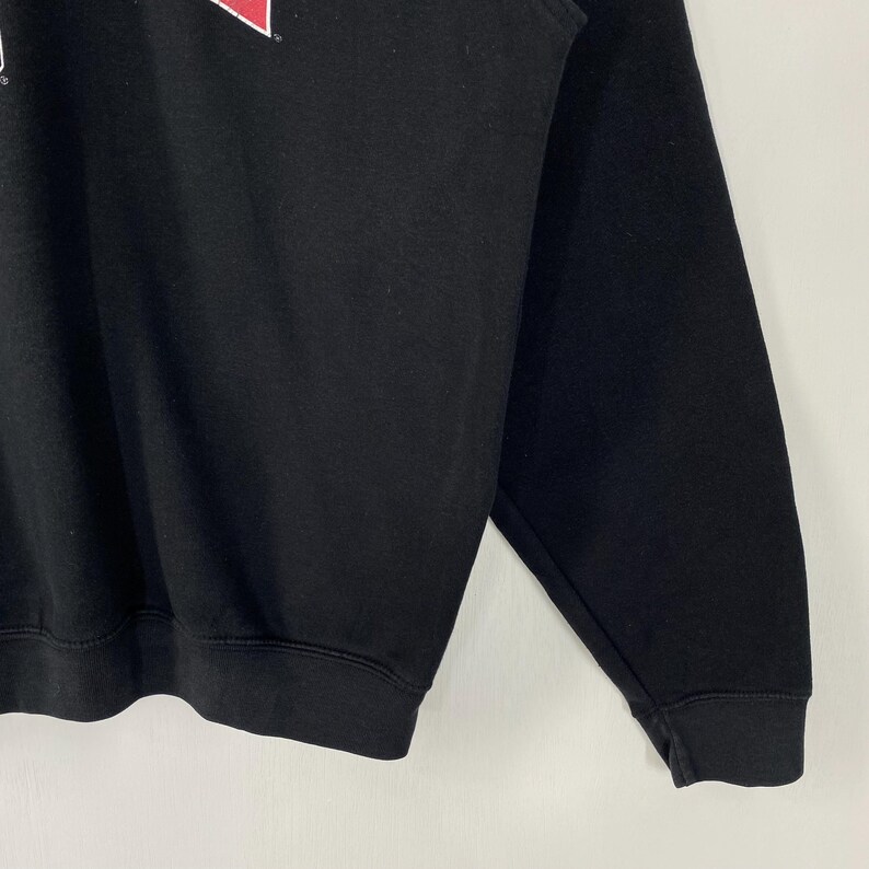 May include: Black sweatshirt with a red and white logo on the upper left chest. The long-sleeved top has a crew neck and ribbed cuffs and hem. The garment is made of a soft, comfortable material.
