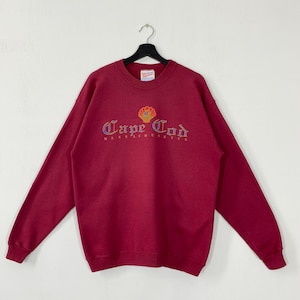 Könnte beinhalten: Ein weinrotes Sweatshirt mit dem gestickten Schriftzug "Cape Cod Massachusetts" in einer verzierten Schriftart, verziert mit einem Muschelmotiv. Das Sweatshirt hat einen Rundhalsausschnitt und lange Ärmel. Es hängt an einem schwarzen Kleiderbügel.