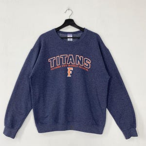 Könnte beinhalten: Ein marineblaues Sweatshirt mit dem Wort "TITANS" in orangefarbenen und weißen Buchstaben auf der Brust. Darunter steht der Text "CALIFORNIA STATE UNIVERSITY FULLERTON" in Orange. Ein orangefarbenes "F" befindet sich darunter. Das Sweatshirt hängt.