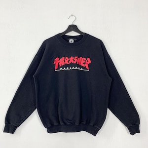 May include: Black sweatshirt with the Thrasher Magazine logo in red and white across the chest. The sweatshirt is hanging on a black hanger against a white background. The long-sleeved top has a crew neck.