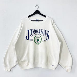 Könnte beinhalten: Cremefarbenes Sweatshirt mit Rundhalsausschnitt und dem Text "JOHNSON & WALES UNIVERSITY" in Marineblau und Grün. Das Sweatshirt hat lange Ärmel und einen gerippten Ausschnitt. Das Universitätslogo befindet sich in der Mitte.