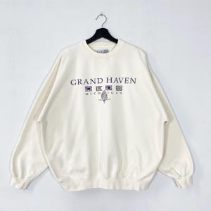 Può includere: Felpa color crema con la scritta "GRAND HAVEN MICHIGAN" in un design multicolore. La felpa ha un girocollo e maniche lunghe ed è appesa a una gruccia nera.