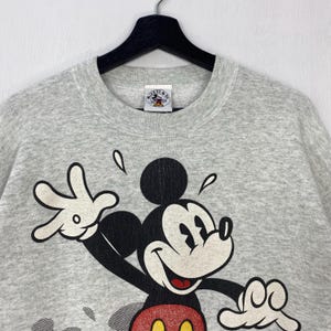 Może przedstawiać: Jasnoszara bluza z dużym nadrukiem Myszki Miki. Myszka Miki jest przedstawiona z białą twarzą, czarnymi uszami i czerwono-ż&oacute;łtym strojem. Bluza ma okrągły dekolt i małą metkę w pobliżu kołnierza.