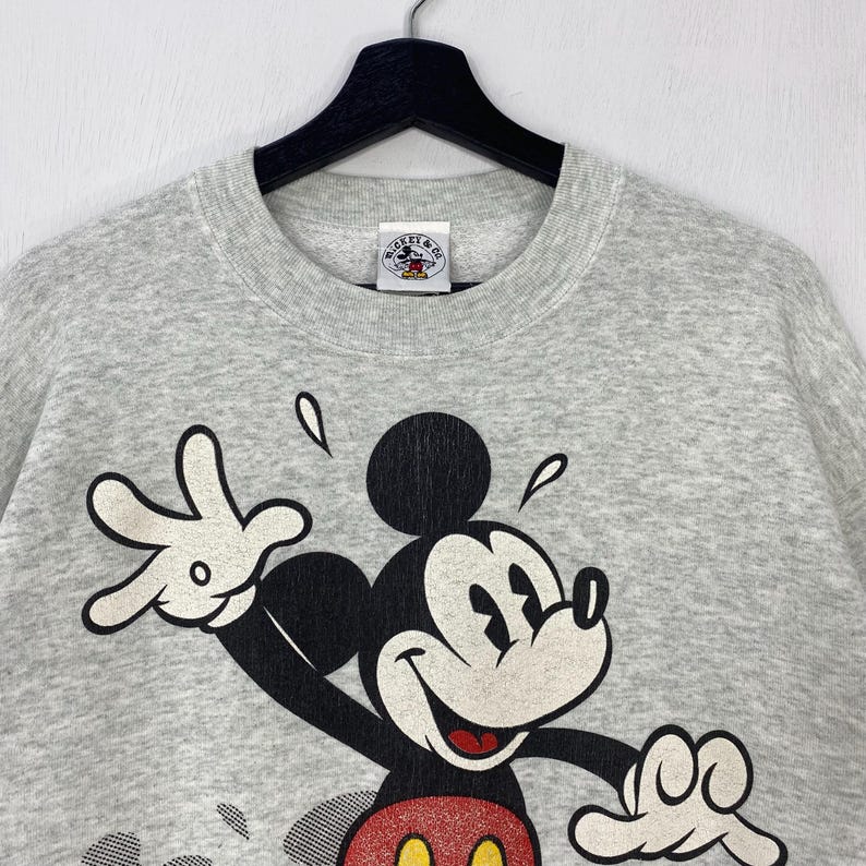 Może przedstawiać: Jasnoszara bluza z dużym nadrukiem Myszki Miki. Myszka Miki jest przedstawiona z białą twarzą, czarnymi uszami i czerwono-ż&oacute;łtym strojem. Bluza ma okrągły dekolt i małą metkę w pobliżu kołnierza.