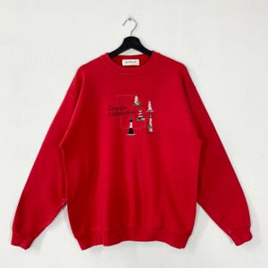 Könnte beinhalten: Ein leuchtend rotes Langarm-Sweatshirt mit Rundhalsausschnitt, das eine gestickte Grafik des Bundesstaates Georgia mit vier Leuchtturm-Illustrationen und dem Text Georgia Lighthouses zeigt. Das Kleidungsstück hängt an einem schwarzen Kleiderbügel.