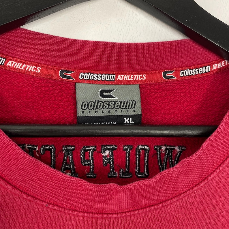K&ouml;nnte beinhalten: Ein rotes Colosseum Athletics XL Sweatshirt. Das Etikett lautet "Colosseum Athletics" und "XL". Das Wort "ATHLETICS" ist ebenfalls schwarz auf der Vorderseite des Sweatshirts gestickt.