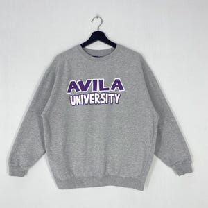 Könnte beinhalten: Ein graues Avila University Sweatshirt mit dem Universitätsnamen in lila Blockbuchstaben. Das Sweatshirt hängt an einem schwarzen Kleiderbügel vor einem weißen Hintergrund. Es hat lange Ärmel und einen Rundhalsausschnitt.