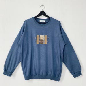 May include: Blue sweatshirt with long sleeves and a crew neck. The front features the text "VENT VERT par PIERRE BALMAIN" in a rectangular design with tan and brown accents. The sweatshirt is hanging on a black hanger.