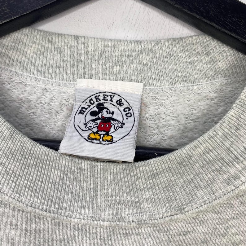 Może przedstawiać: Zbliżenie szarej bluzy z metką "Mickey & Co.". Etykieta przedstawia Myszka Miki w swoim klasycznym stroju w okrągłym wzorze. Bluza ma okrągły dekolt i teksturowaną tkaninę.