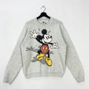 Może przedstawiać: Jasnoszara bluza z dużym nadrukiem Myszki Miki. Myszka Miki jest przedstawiona w dynamicznej pozie, z wyciągniętymi ramionami i wesołym wyrazem twarzy. Ma na sobie charakterystyczne czerwone spodenki i ż&oacute;łte buty.