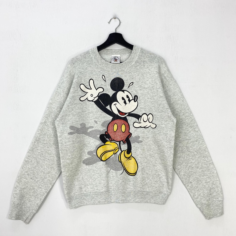 Może przedstawiać: Jasnoszara bluza z dużym nadrukiem Myszki Miki. Myszka Miki jest przedstawiona w dynamicznej pozie, z wyciągniętymi ramionami i wesołym wyrazem twarzy. Ma na sobie charakterystyczne czerwone spodenki i ż&oacute;łte buty.