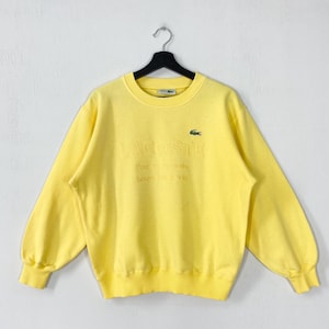 Op de afbeelding: Een gele Lacoste sweatshirt met de merknaam en het logo. De sweatshirt heeft een ronde hals en lange mouwen. De tekst "Pour les moments heureux de la vie" staat eronder.