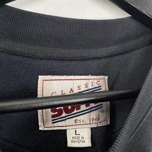 May include: Close-up of a navy blue crew neck shirt. The shirt features a tag with the words "CLASSIC" and "SUPP" in red and white. The tag also reads "Est. 1946" and "L Made in Pakistan".