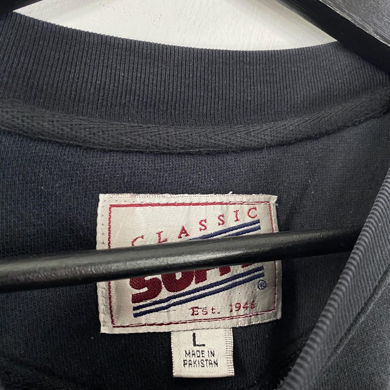 May include: Close-up of a navy blue crew neck shirt. The shirt features a tag with the words "CLASSIC" and "SUPP" in red and white. The tag also reads "Est. 1946" and "L Made in Pakistan".