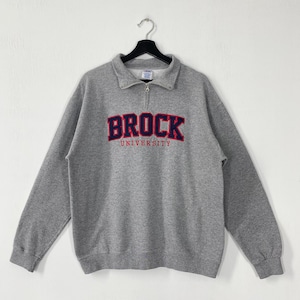 May include: A grey sweatshirt with a collared neckline and a quarter-zip. The word "BROCK" is printed across the chest in red and navy blue, with "UNIVERSITY" below. The sweatshirt is hanging on a black hanger.