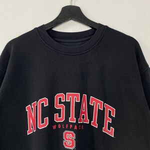 May include: Black sweatshirt with the words "NC STATE" in red block letters with a white outline. Below the text is the word "WOLFPACK" in smaller red letters. A red and white "N" and "S" logo is at the bottom.