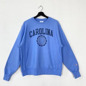 Peut inclure: Sweat-shirt bleu clair avec le mot "CAROLINA" imprim&eacute; en bleu marine sur la poitrine. En dessous, un &eacute;cusson bleu marine. Le logo Champion est visible sur la manche gauche.