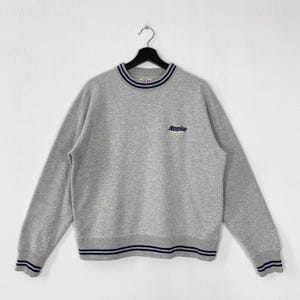 Op de afbeelding: Een grijze sweatshirt met een ronde hals en lange mouwen. De kraag, manchetten en zoom hebben marineblauwe strepen. Het woord "RonJon" is in marineblauw op de borst geborduurd. De sweatshirt hangt aan een zwarte hanger.