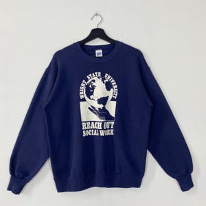 May include: A navy blue sweatshirt featuring the text "WRIGHT STATE UNIVERSITY" and "REACH OUT SOCIAL WORK" in white. The graphic shows a globe held by a hand. The sweatshirt is suspended from a black hanger.