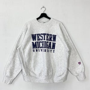Könnte beinhalten: Hellgraues Sweatshirt mit dem navyblauen Schriftzug "WESTERN MICHIGAN UNIVERSITY". Das Sweatshirt hat lange Ärmel und ein kleines Emblem auf dem linken Ärmel. Es hängt an einem schwarzen Kleiderbügel.