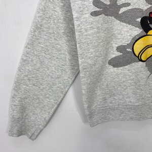 Może przedstawiać: Jasnoszara bluza z postacią z kresk&oacute;wki z ż&oacute;łtymi rękawicami i czarno-czerwoną twarzą. Postać jest osadzona na szarym, kropkowanym tle. Bluza ma długie rękawy i okrągły dekolt.