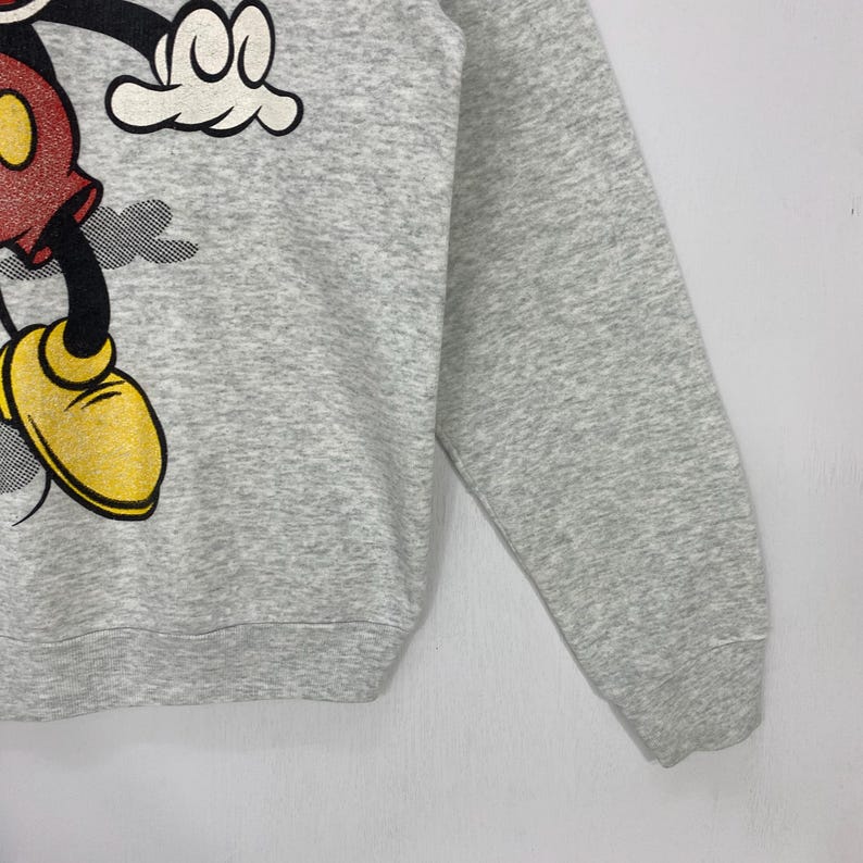 Może przedstawiać: Szara bluza z grafiką Myszki Miki. Postać z kresk&oacute;wki jest przedstawiona w swoim klasycznym stroju z czerwonymi spodenkami, ż&oacute;łtymi butami i białymi rękawiczkami. Bluza ma okrągły dekolt i długie rękawy.