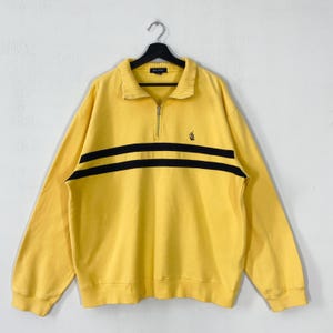 Puede incluir: Sudadera amarilla con dos rayas horizontales negras. Tiene cuello con cremallera y un pequeño logotipo. La marca "NAUTICA" es visible en el cuello. La parte superior de manga larga está colgada de una percha negra.