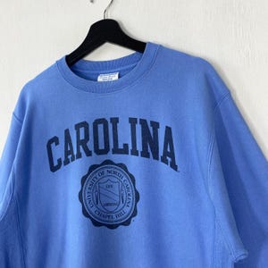 Peut inclure: Un sweat-shirt bleu clair avec le mot "CAROLINA" en bleu marine en arc sur la poitrine. En dessous, un &eacute;cusson circulaire bleu marine avec le texte "UNIVERSITY OF NORTH CAROLINA CHAPEL HILL". Le sweat-shirt est suspendu &agrave; un cintre noir.