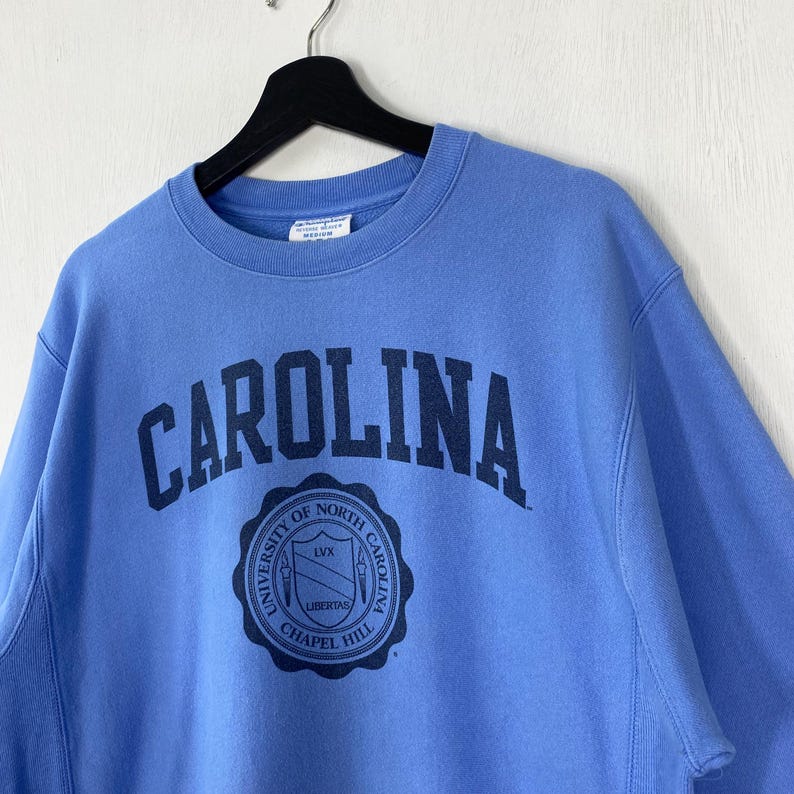 Peut inclure: Un sweat-shirt bleu clair avec le mot "CAROLINA" en bleu marine en arc sur la poitrine. En dessous, un &eacute;cusson circulaire bleu marine avec le texte "UNIVERSITY OF NORTH CAROLINA CHAPEL HILL". Le sweat-shirt est suspendu &agrave; un cintre noir.