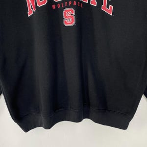 May include: Black sweatshirt with the text "WOLFPACK" in red block letters across the top. Below the text is a red and white logo with the letter "S" inside a red and white outline of the letters "NC".