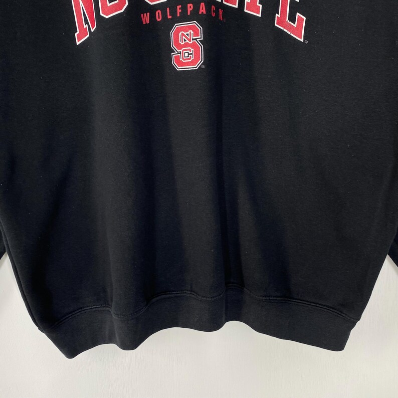 May include: Black sweatshirt with the text "WOLFPACK" in red block letters across the top. Below the text is a red and white logo with the letter "S" inside a red and white outline of the letters "NC".